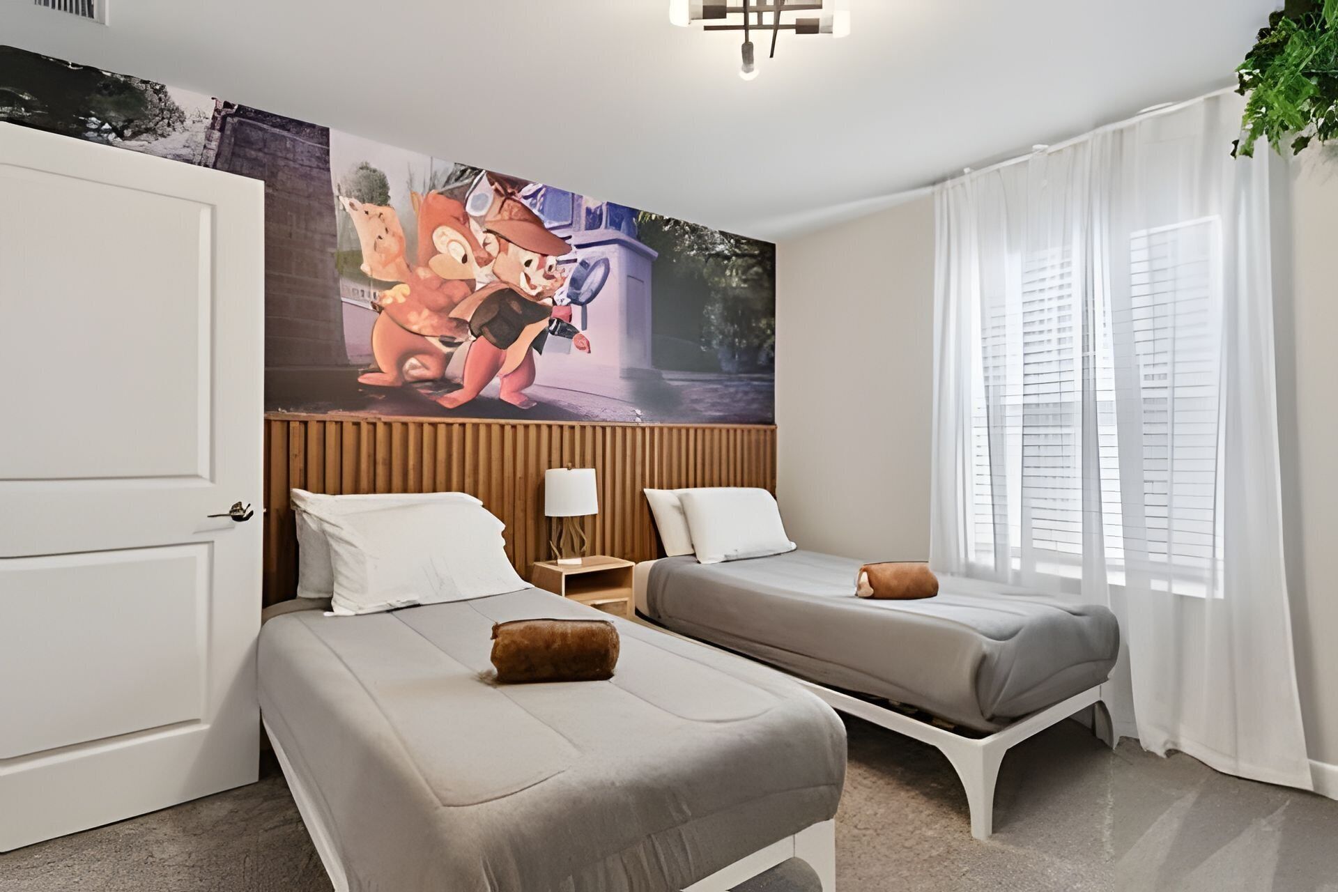 A hotel room with two single beds, a wooden headboard, a wall mural, a bedside lamp, and a window with sheer curtains.