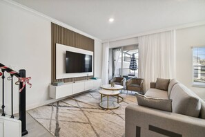 A modern living room with a flat-screen TV mounted on a white wall, a beige sofa, a small round coffee table, and a patterned rug. - Storey Lake 5bd/4ba Private Pool Near Disney (Kissimmee)