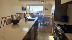 Une cuisine moderne dotée d’un îlot central, d’un coin repas avec des chaises, d’une machine à café et offrant une vue sur la mer grâce à de grandes fenêtres.