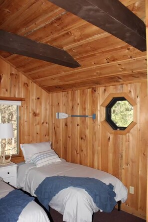 A wooden cabin room with two beds, a window, and a wall-mounted lamp.