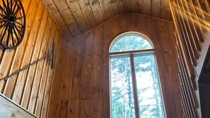 Een chaletinterieur met houten wanden, een schuine balkenlaag, een plafondventilator, een raam met uitzicht op bomen, en een rustieke houten deur.