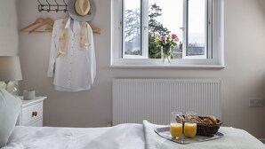 Ein Schlafzimmer mit Bett, Nachttisch, Fenster mit Blick auf Bäume, Heizkörper und Tablett mit Croissants und Saft.