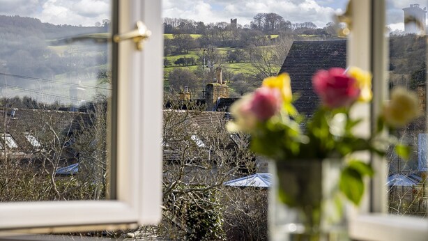 Blick durch ein Fenster, im Vordergrund eine Blumenvase, eine ländliche Landschaft mit einem Turm in der Ferne und einem klaren Himmel.