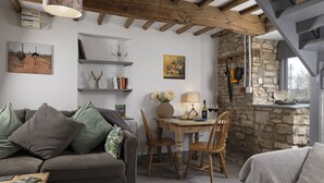 Ein gemütliches Wohnzimmer mit Holzbalkendecke, Steinwand, Esstisch mit Stühlen und Bücherregal mit Büchern und Dekoration.