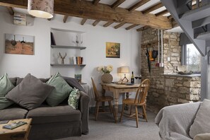 Ein gemütliches Wohnzimmer mit Holzbalkendecke, Steinwand, Esstisch mit Stühlen und Bücherregal mit Büchern und Dekoration.