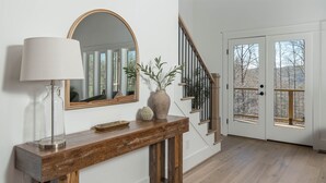 A wooden console table with a lamp, a mirror, and decorative items. A staircase with a wooden railing leads to a door with glass panels. - Luxury MTN Retreat | Hot Tub | Fire Pit | Game Rm (Fleetwood)