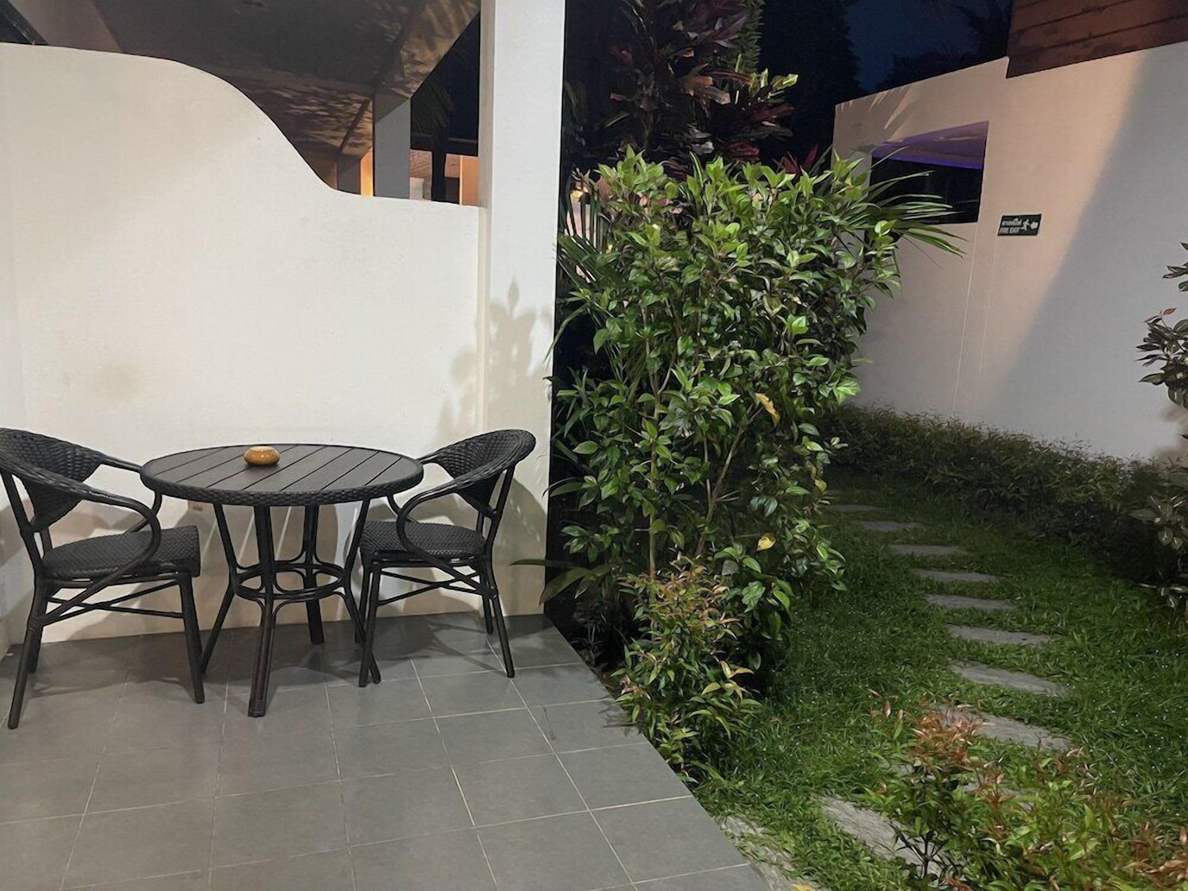 A patio with a round table and four chairs, surrounded by a white wall and a small garden with a hedge.