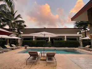 A pool area with lounge chairs and umbrellas, surrounded by palm trees and a building with a flat roof. - Lotus Bleu Resort & Restaurant (Rawai)