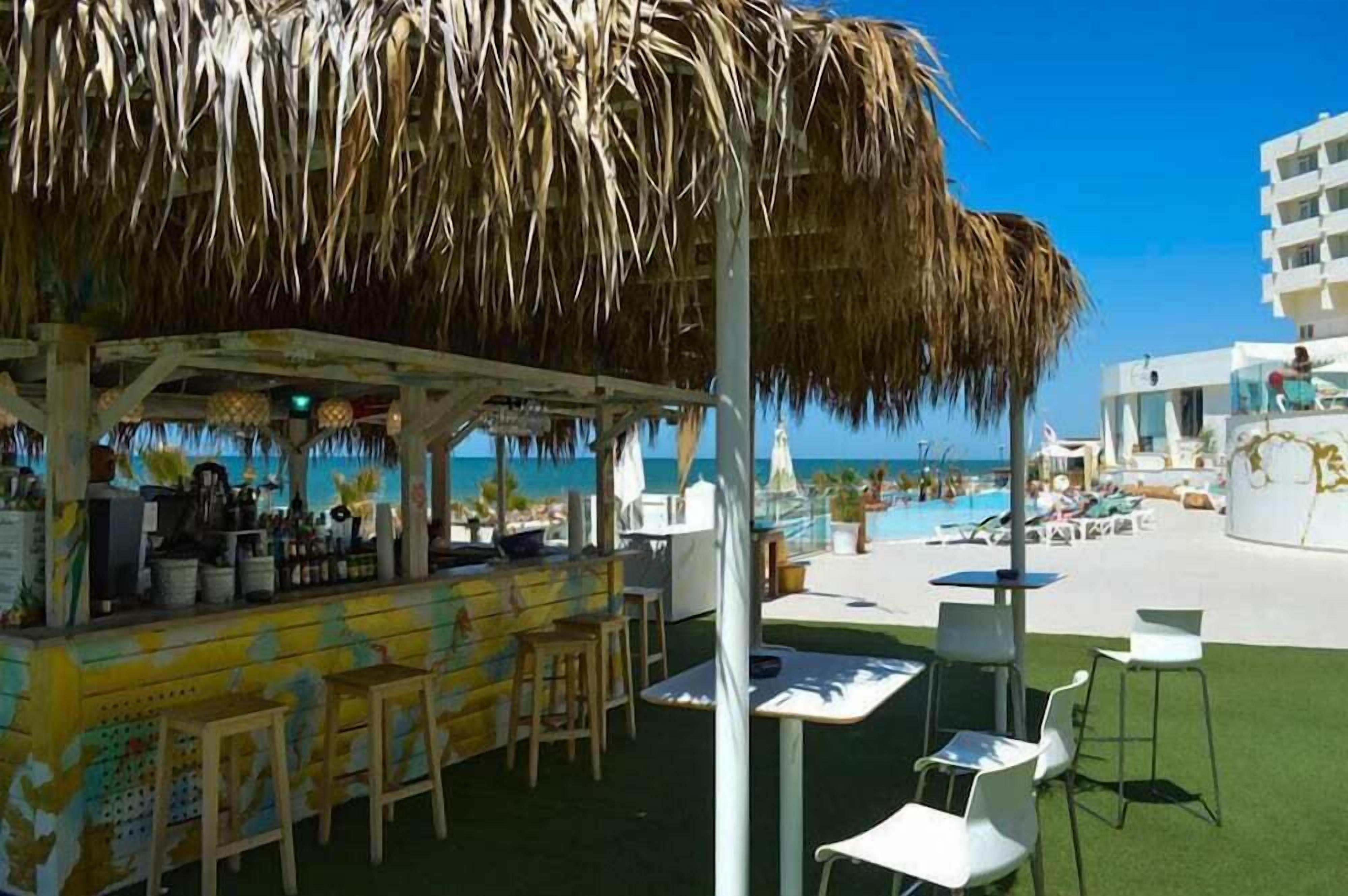 Un bar en la playa con techo de paja, mesas al aire libre y vistas al océano.