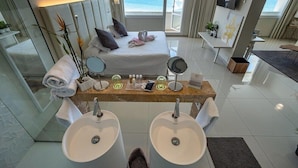 A modern hotel room with a bed, two sinks, a vanity, and a large window overlooking the ocean. - On Hotels Oceanfront Adults Designed (Almonte)