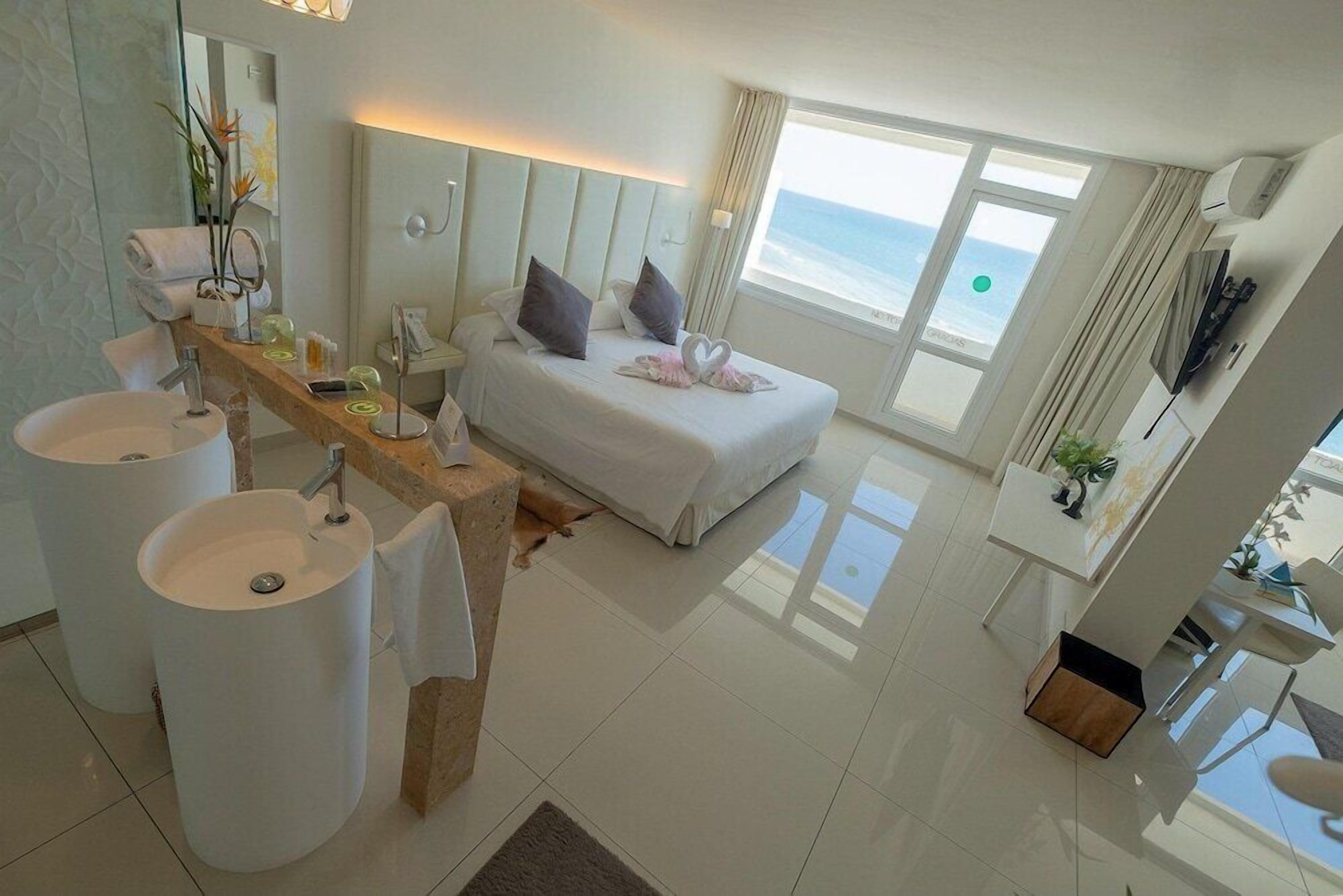Una habitación de hotel moderna con una cama grande, dos lavamanos blancos, un tocador con espejo y vista al océano.