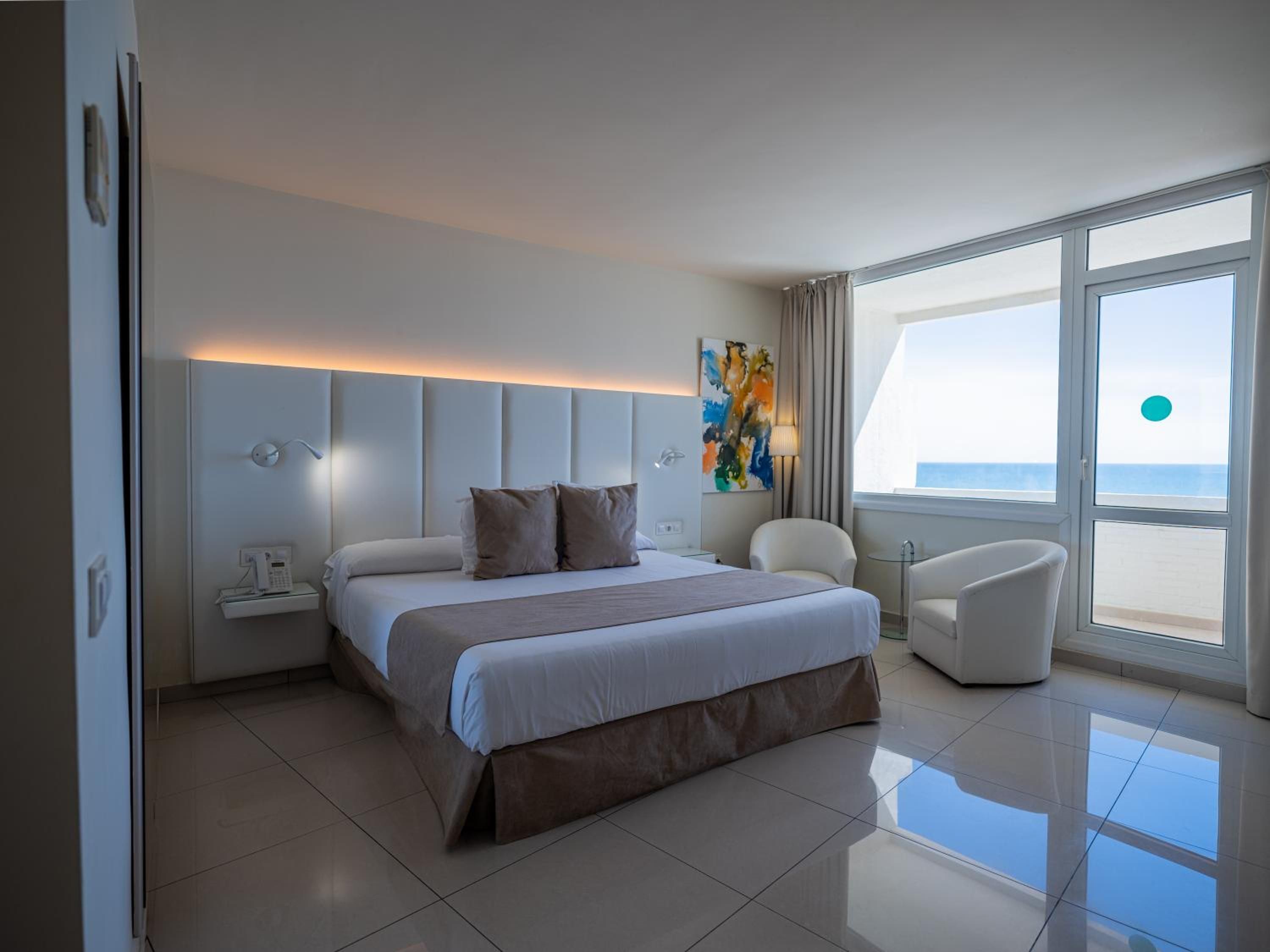 Habitación de hotel moderna con una cama grande, una mesita de noche con teléfono, una silla y vista al mar.