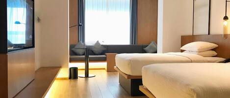 Ein modernes Hotelzimmer mit zwei Betten, einer Couch, einem Fernseher und einem großen Fenster mit Vorhängen.