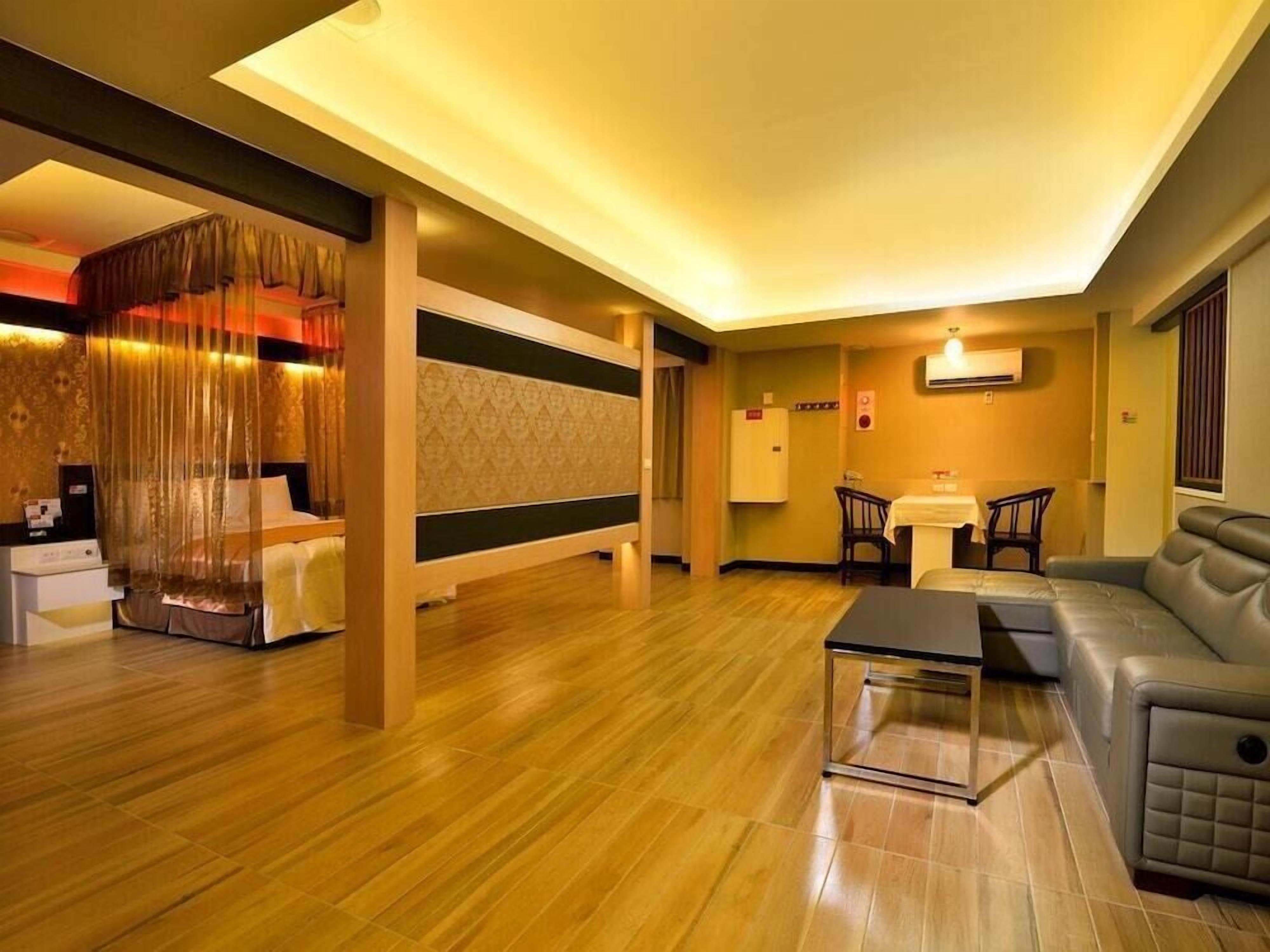 Une chambre d’hôtel avec un plancher en bois, un canapé, une petite table, un lit avec une tête de lit, un rideau et un climatiseur fixé au mur.