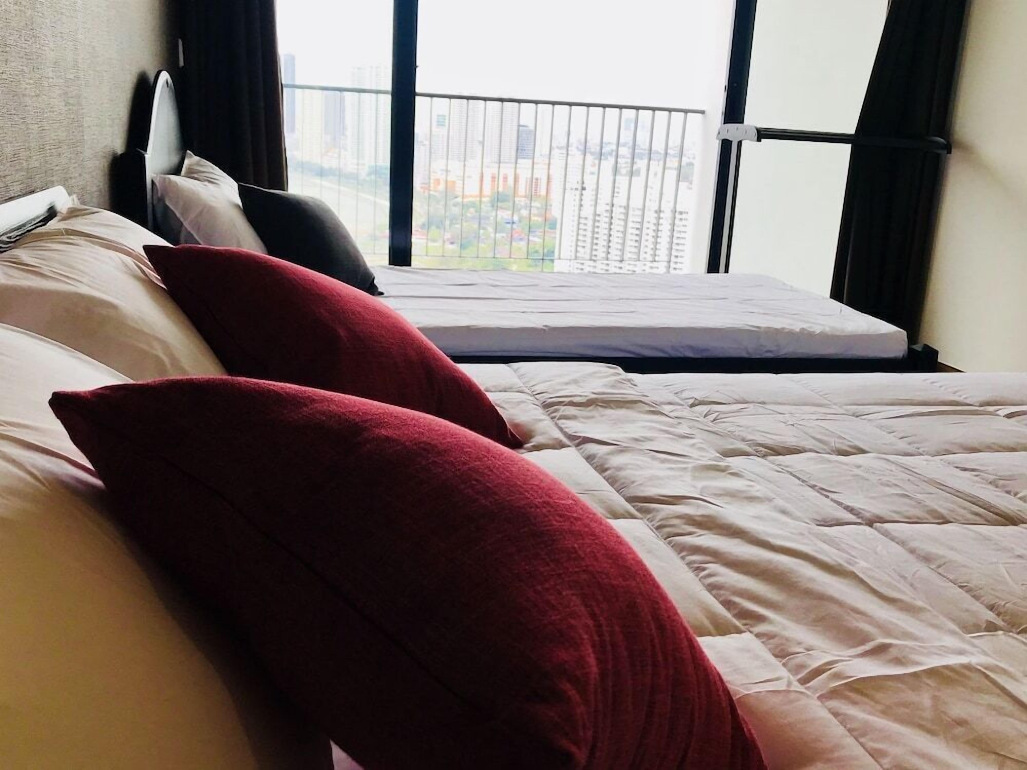 A hotel room with a bed, two red pillows, a white mattress, and a view of a cityscape through a window with a black metal railing.