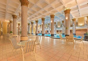 Ett rymligt inomhusområde med en pool, omgivet av pelare med intrikata mönster, och inrett med randiga stolar och bord.