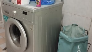 A front-loading washing machine with a control panel, a laundry bag labeled "LAUNDRY" hanging nearby, and a tiled floor.