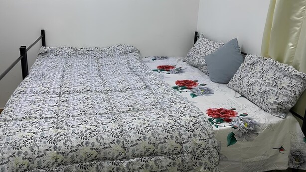 A single bed with a floral-patterned bedspread and pillows, a plain wall, and a curtain on the right side.