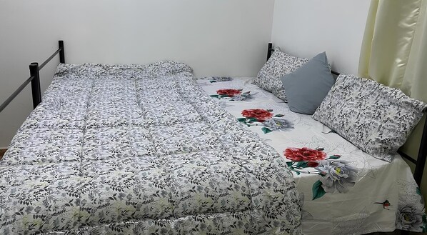 Un lit simple avec une couvre-lit et des oreillers à motifs floraux, un mur uni et un rideau sur le côté droit.