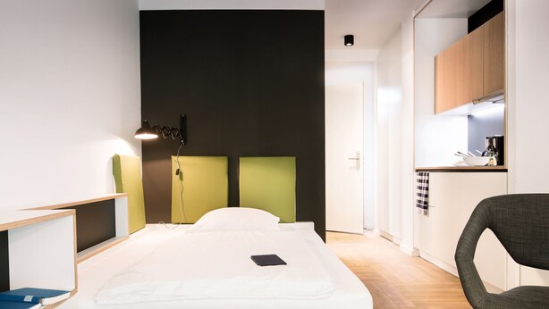A modern hotel room with a bed, bedside table, lamp, and a small kitchenette area. - Smartment Business Berlin Karlshorst (Berlin)
