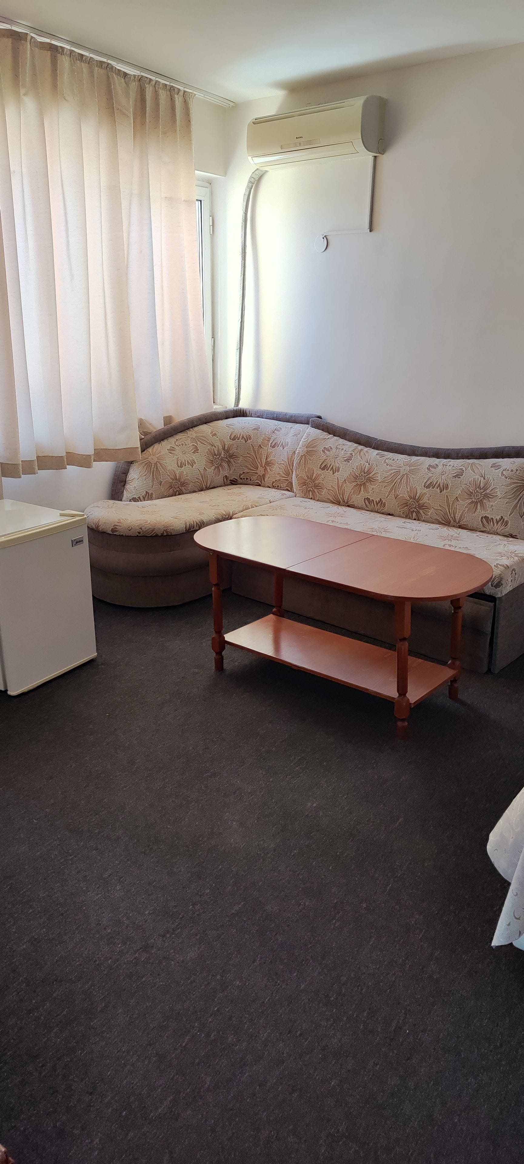 A living room with a sofa, a small table, a refrigerator, and an air conditioning unit on the wall.