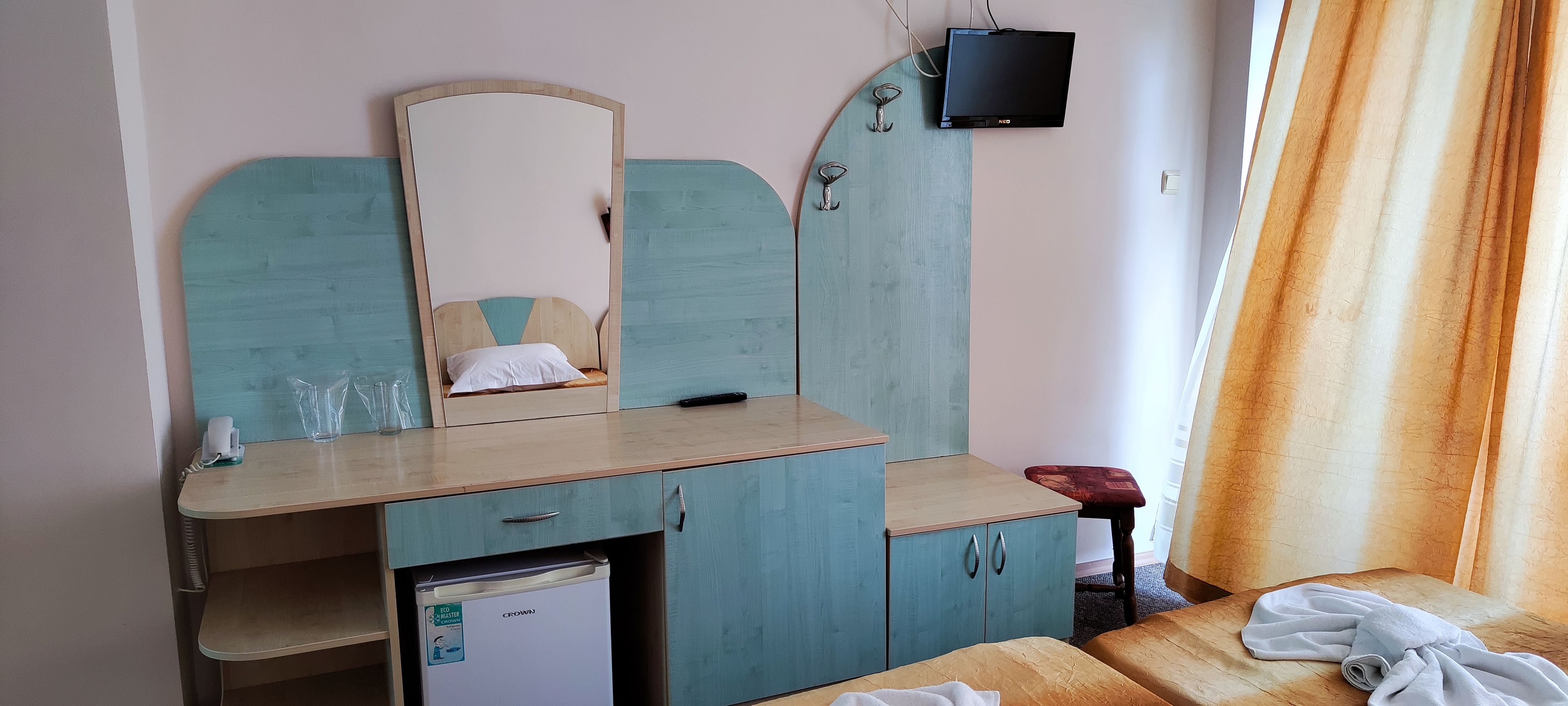 A bedroom with a bed, a small refrigerator, a wooden desk with a mirror, and a TV mounted on the wall.