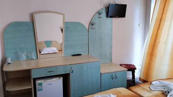 A bedroom with a bed, a small refrigerator, a wooden desk with a mirror, and a TV mounted on the wall.