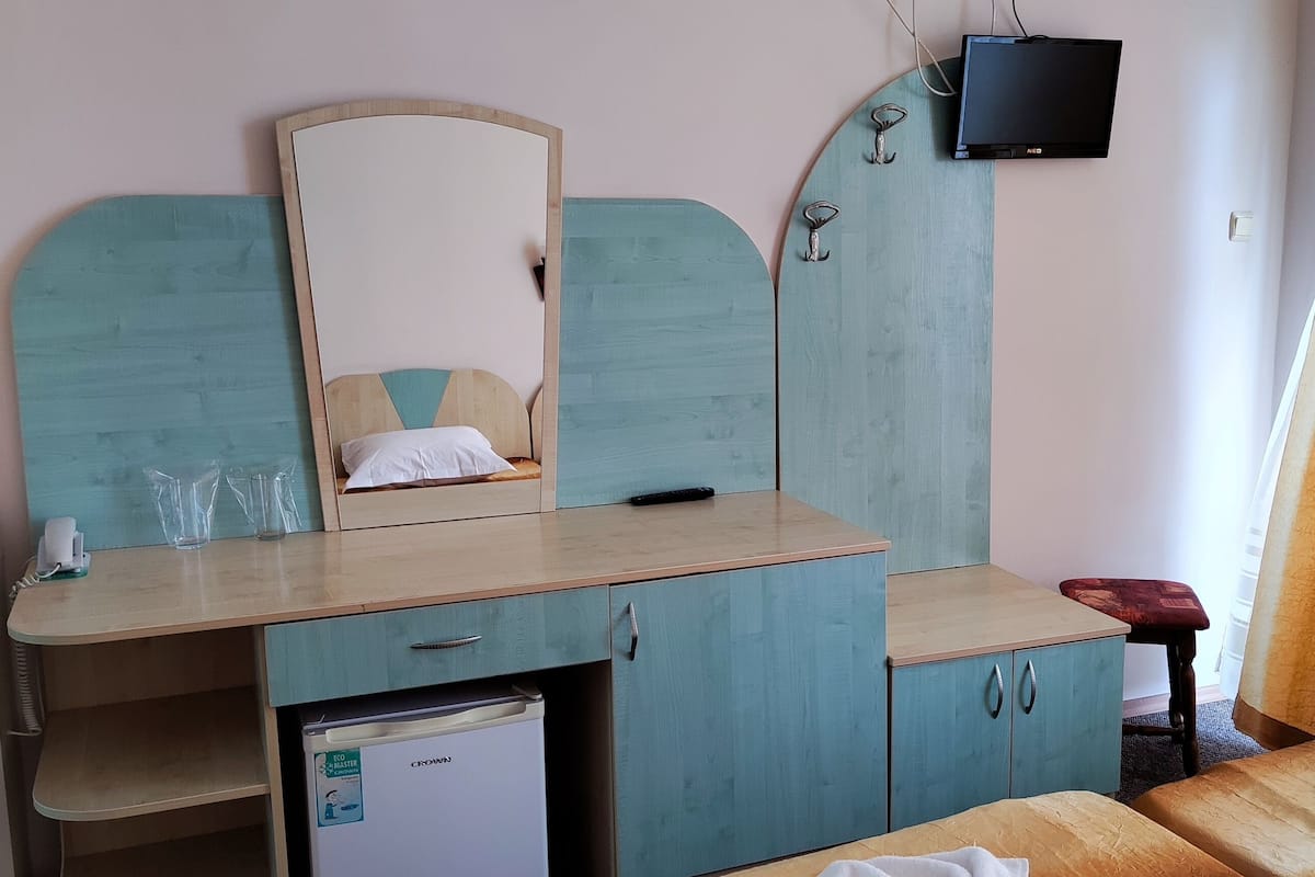 A bedroom with a bed, a small refrigerator, a wooden desk with a mirror, and a TV mounted on the wall.