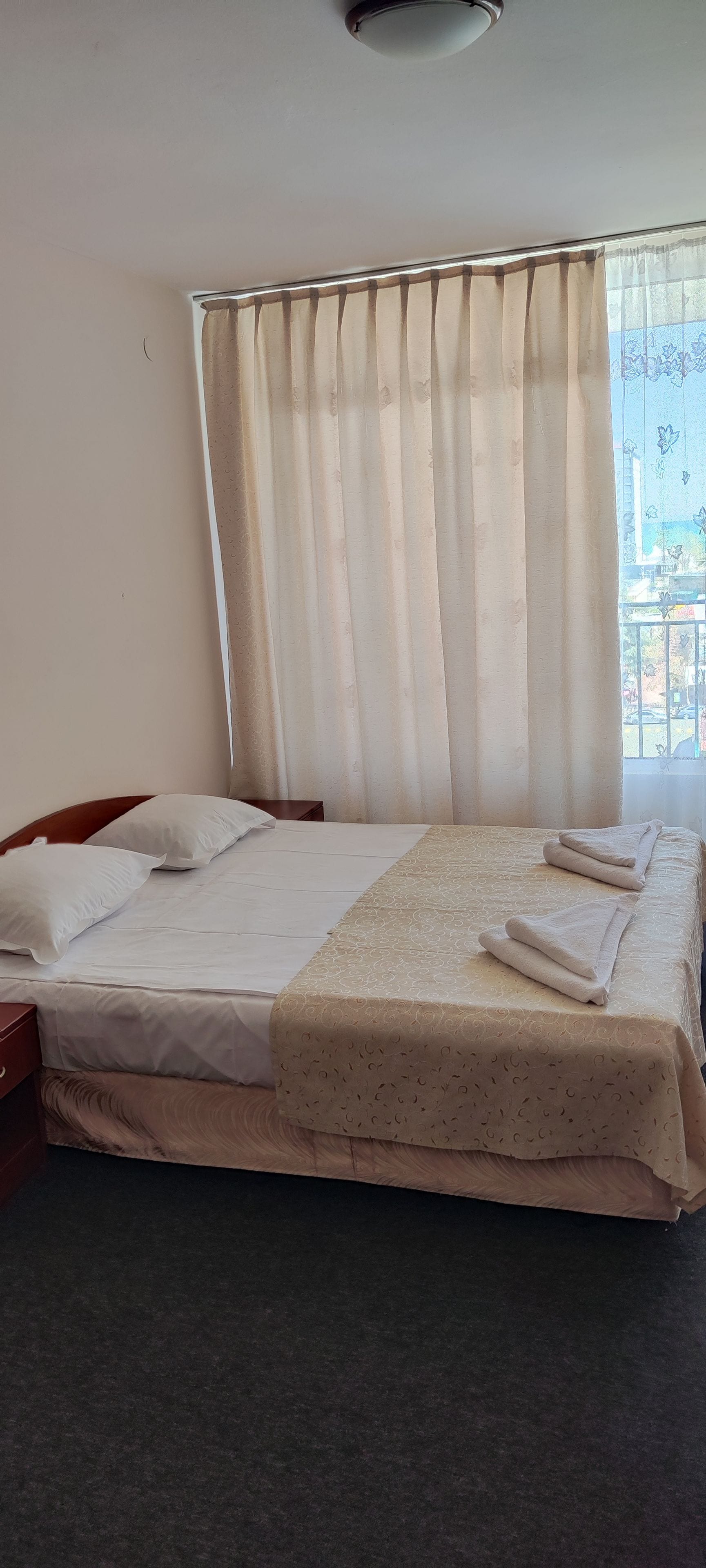 A neatly made bed with a patterned bedspread, a window with curtains, and a view of a balcony.