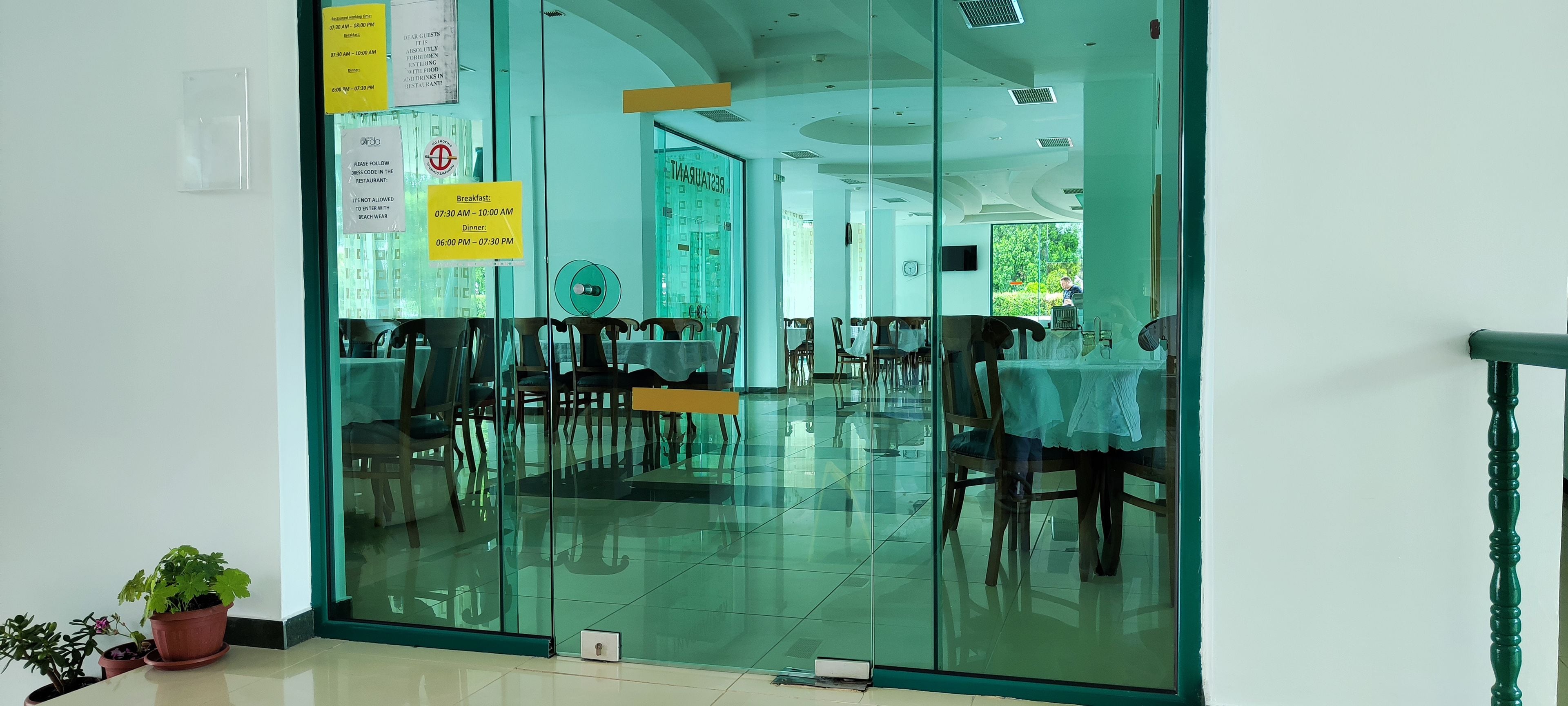 A restaurant with a glass partition, wooden chairs, and tables. There are potted plants on the floor.