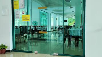 A restaurant with a glass partition, wooden chairs, and tables. There are potted plants on the floor.