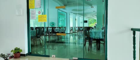 Un restaurante con separador de vidrio, sillas y mesas de madera. Hay plantas en macetas en el suelo.
