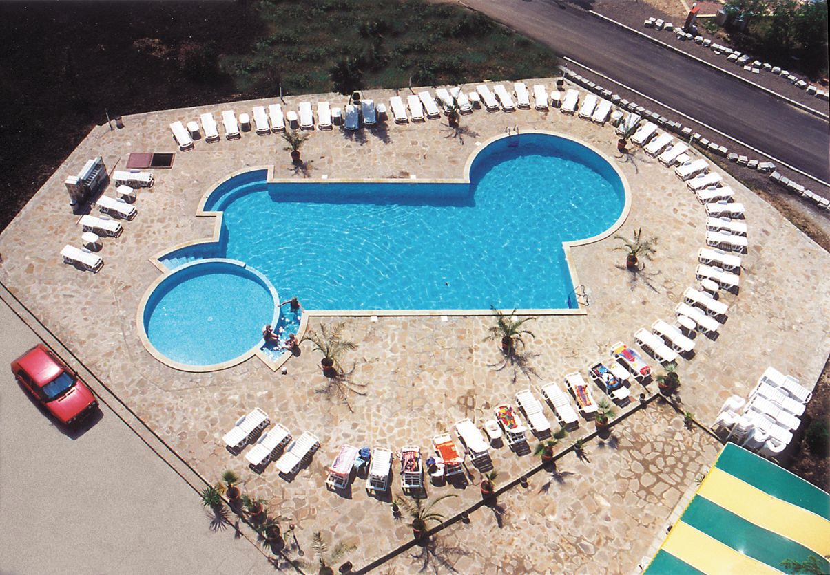 Una piscina con forma de coche, rodeada de sillones de descanso y un coche rojo estacionado cerca.