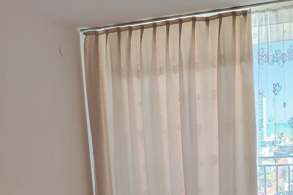 A neatly made bed with a patterned bedspread, a window with curtains, and a view of a balcony.