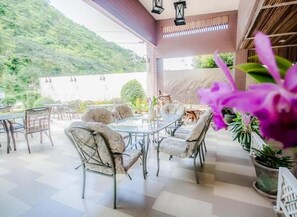Une terrasse extérieure avec un espace repas comprenant des tables et des chaises, entourée de plantes en pot et offrant une vue sur une colline.