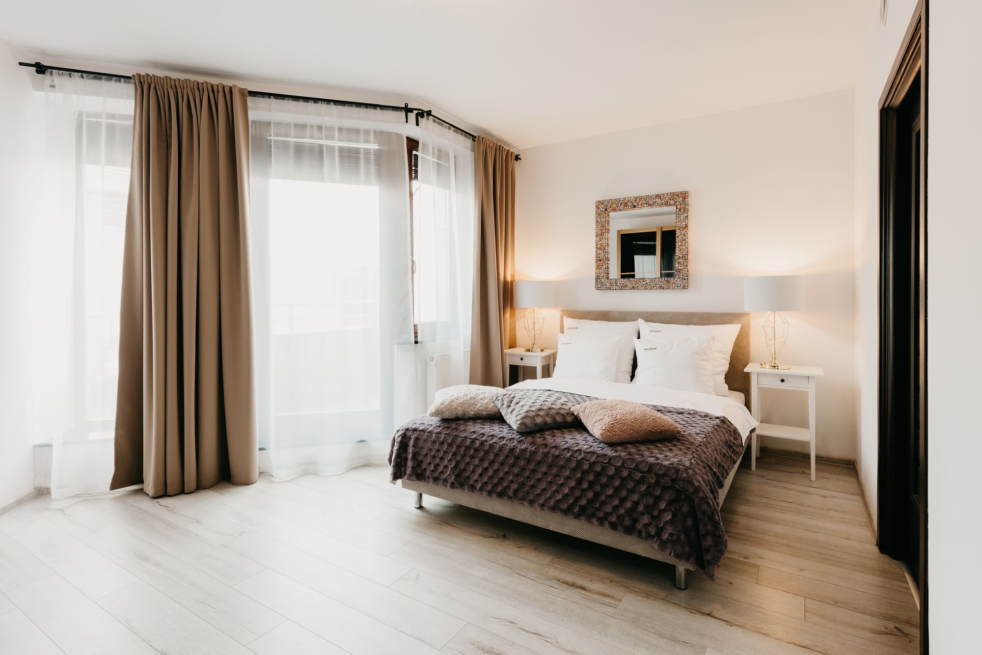 A modern bedroom with a large bed, a nightstand with a lamp, a mirror with a decorative frame, and sheer curtains.