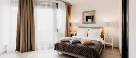 Un dormitorio moderno con una cama grande, una mesita de noche con lámpara, un espejo con marco decorativo y cortinas traslúcidas.