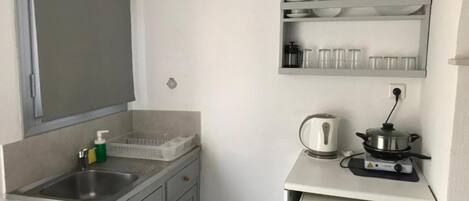 A compact kitchenette with a sink, a kettle, a small stove, and a shelf with dishes and glasses.