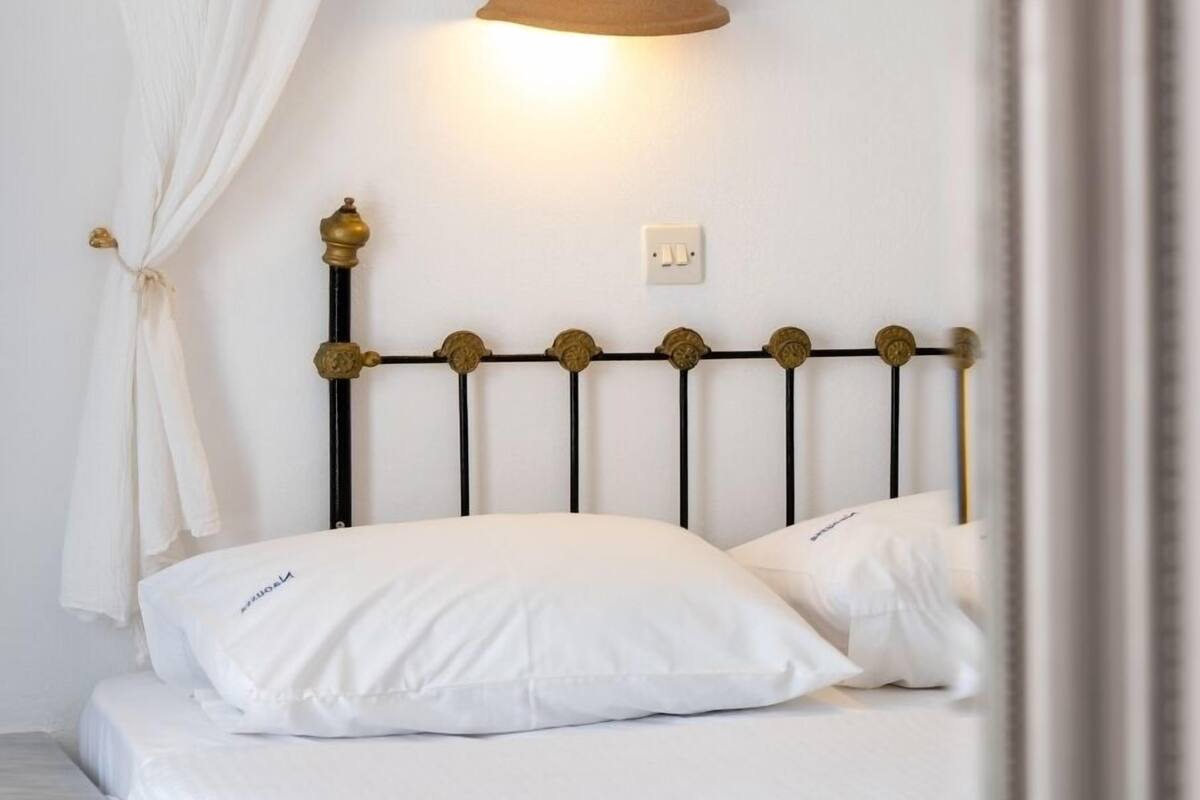 Una cama con cabecera, una lámpara de pared y un espejo que refleja parte de la habitación.