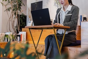 A person sitting on a wooden bench with a laptop, surrounded by books and plants. - H Daimyo (Fukuoka)