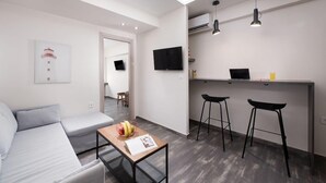 A modern living room with a sofa, a coffee table with fruits, and a TV on the wall. There is a kitchen area with a bar and stools. - Xenofontos Inn by Livin Urbban (Athens)