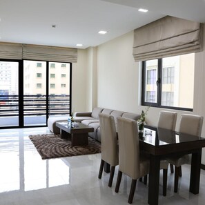 A modern living room with a sofa, a dining table with chairs, a coffee table, and a large window with a view of buildings. - Belle Tower Luxury Hotel Apartments (Manama)