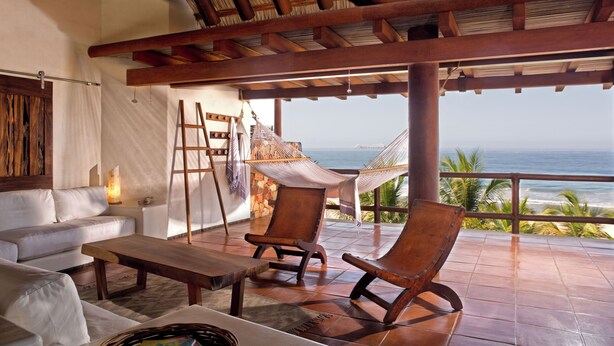 Habitación frente a la playa con techo de madera, una hamaca, una silla de madera, un sofá y vista al océano.