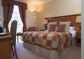 A hotel room with a bed, two bedside tables, a desk with a chair, a lamp, and a window with curtains. - The Bridge Hotel And Spa (Wetherby)