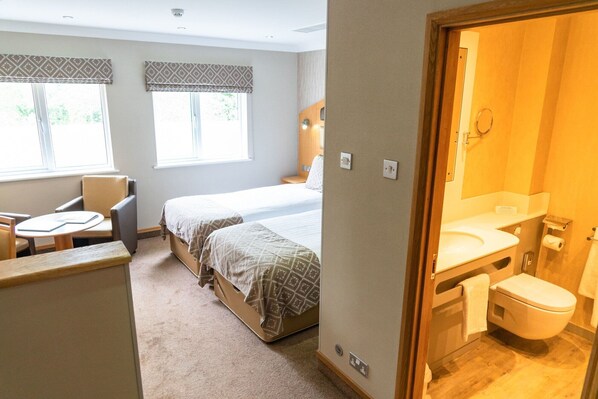 A hotel room with two beds, a small table, a chair, a window with blinds, and a bathroom with a toilet and sink. - The Fairlawns Hotel And Spa (Walsall)