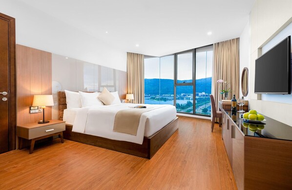 Habitación de hotel con una cama grande, un escritorio con silla, televisión y vistas a un paisaje urbano a través de amplias ventanas.