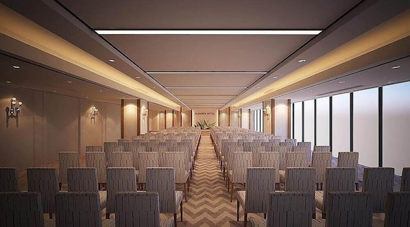 Une salle de conférence d’hôtel avec des rangées de chaises, une allée centrale et de grandes fenêtres sur un côté.