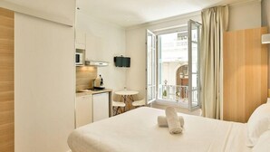 A hotel room with a bed, a small desk with a chair, a TV mounted on the wall, and a window with a view of a building outside. - Nice Home Studio (Nice)