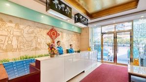 A hotel reception area with a receptionist, a decorative wall with cultural motifs, and a view of the street outside.