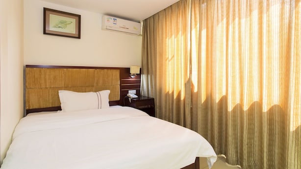 A hotel room with a large bed, a wooden headboard, a bedside table with a lamp, and a window with curtains.